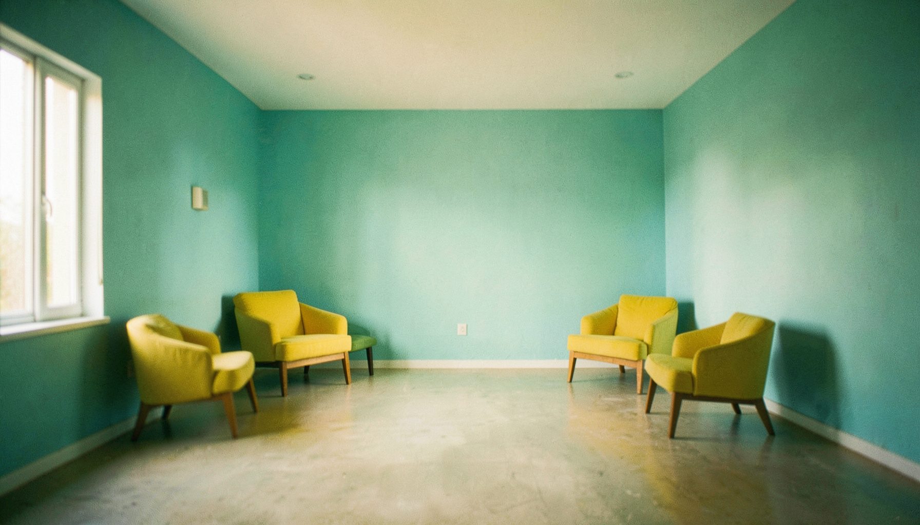 Wide shot of a plain open room with turquoise walls and yellow accent furniture, soft natural light, calm empty atmosphere
